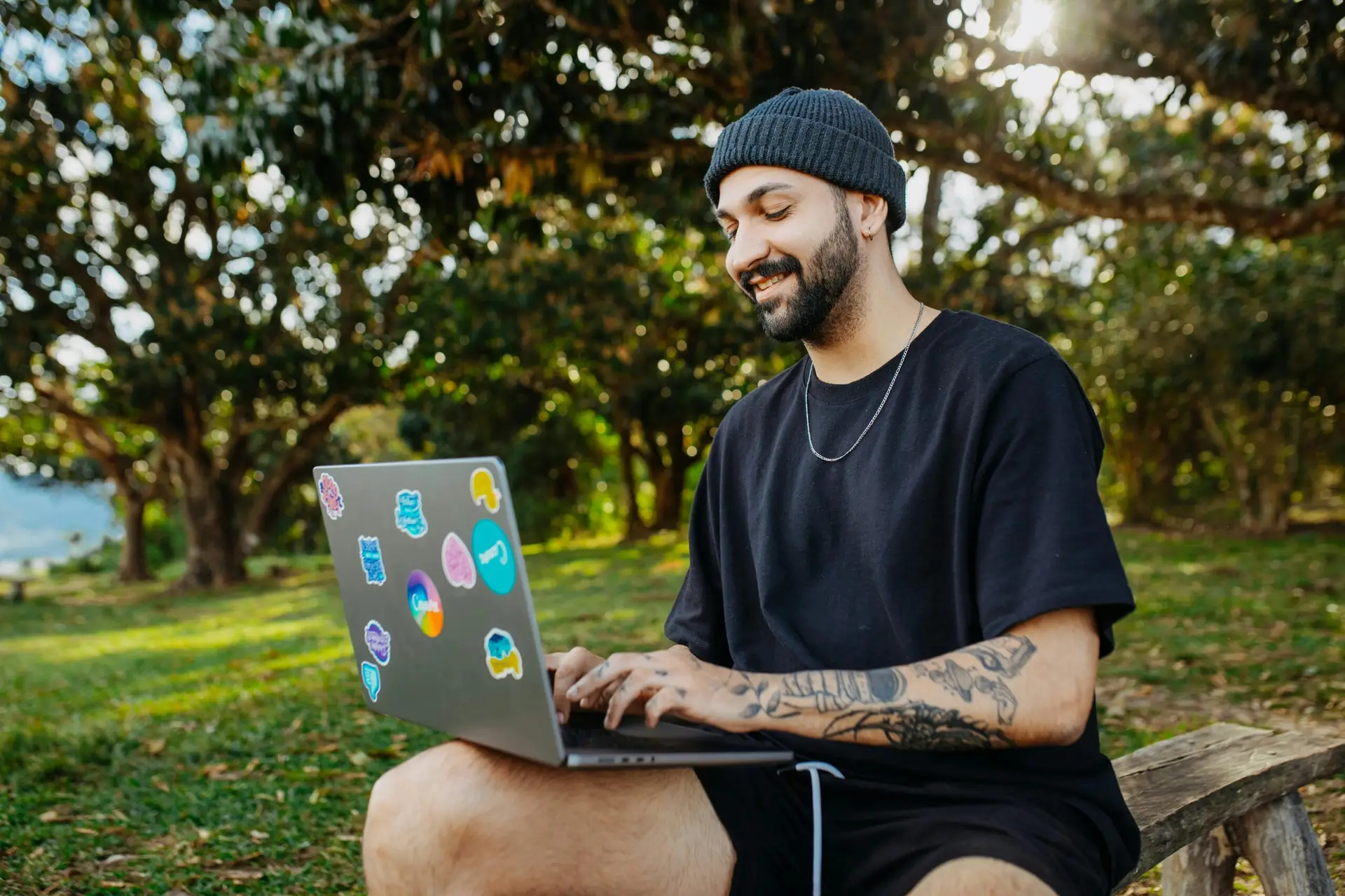 Smiling man with a beard using a laptop outdoors in a serene natural setting.