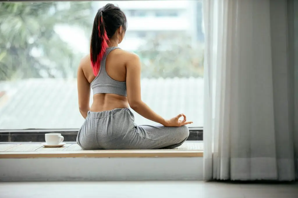 Back view of faceless female with pink hair sitting on mat in Lotus pose with mudra hands and meditating while practicing yoga